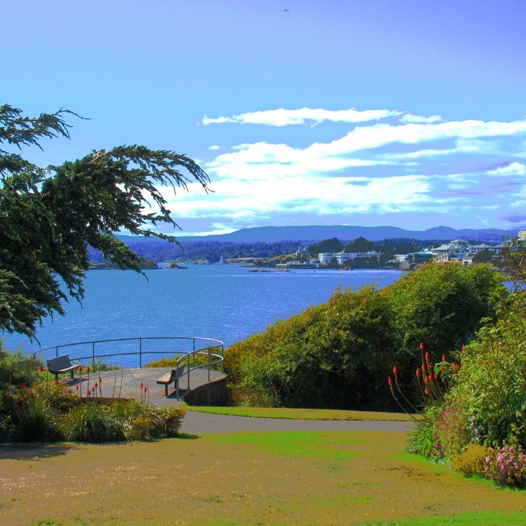 Seaside park with benches on a viewing platform overlooking a calm bay and distant town under a blue sky