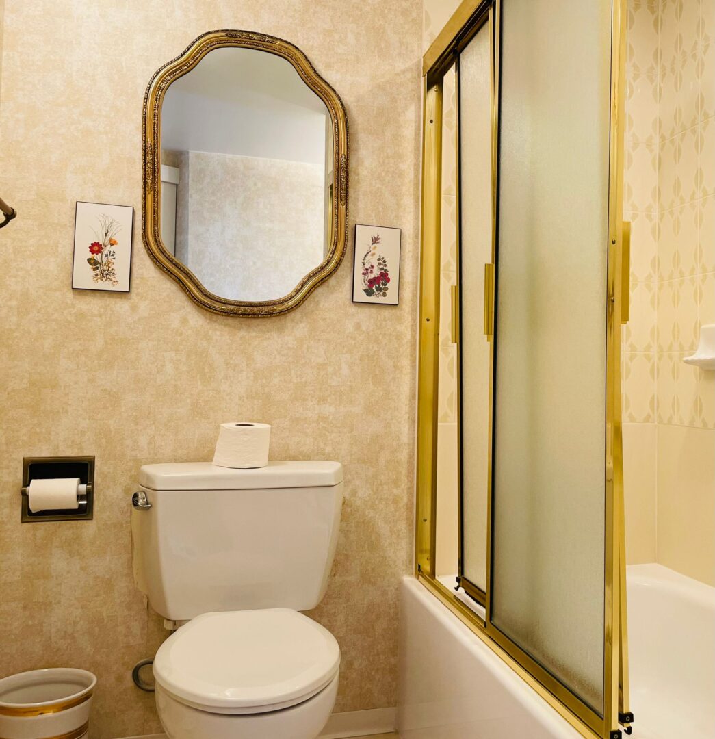 2225belleville10 | Apartments Homes Suites for Rent Victoria BC A vintage style bathroom with a white toilet toilet paper holder a gold ornate mirror and floral art prints on beige walls next to a frosted glass shower enclosure with a brass frame