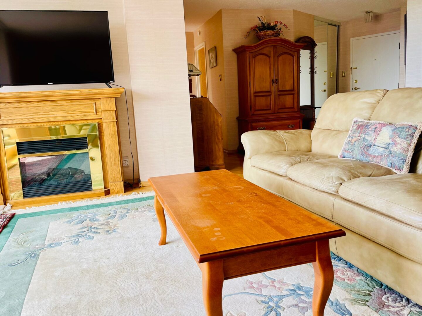 2225belleville5 | Apartments Homes Suites for Rent Victoria BC Living room scene with a cream leather sofa a wooden coffee table and a large flat screen TV on a mantel plus a tall wooden armoire against beige walls