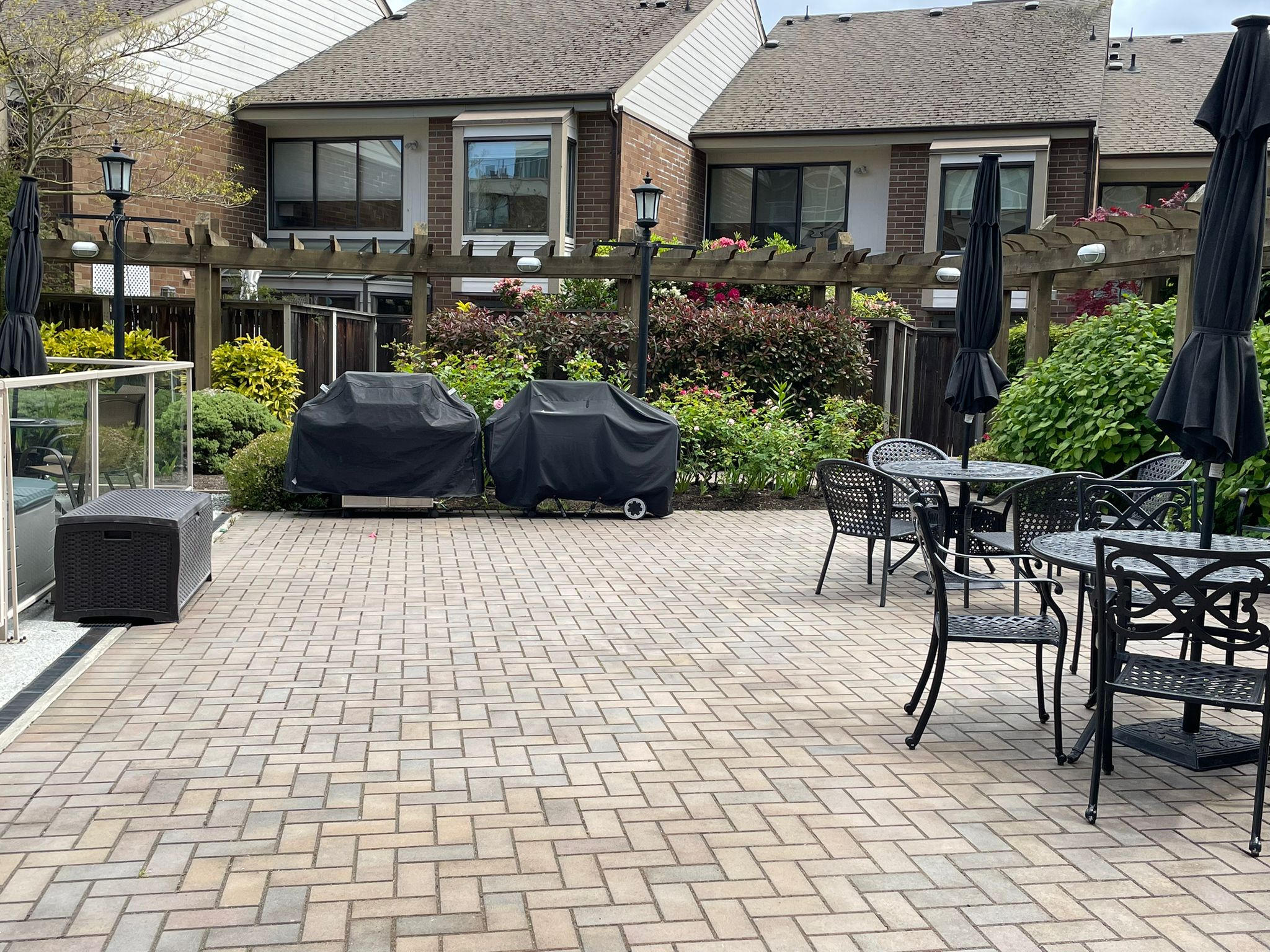 6225belleville16 | Apartments Homes Suites for Rent Victoria BC Backyard patio area with a round metal table and chairs tall black umbrellas grill covers plants and a wooden pergola against brick townhouses
