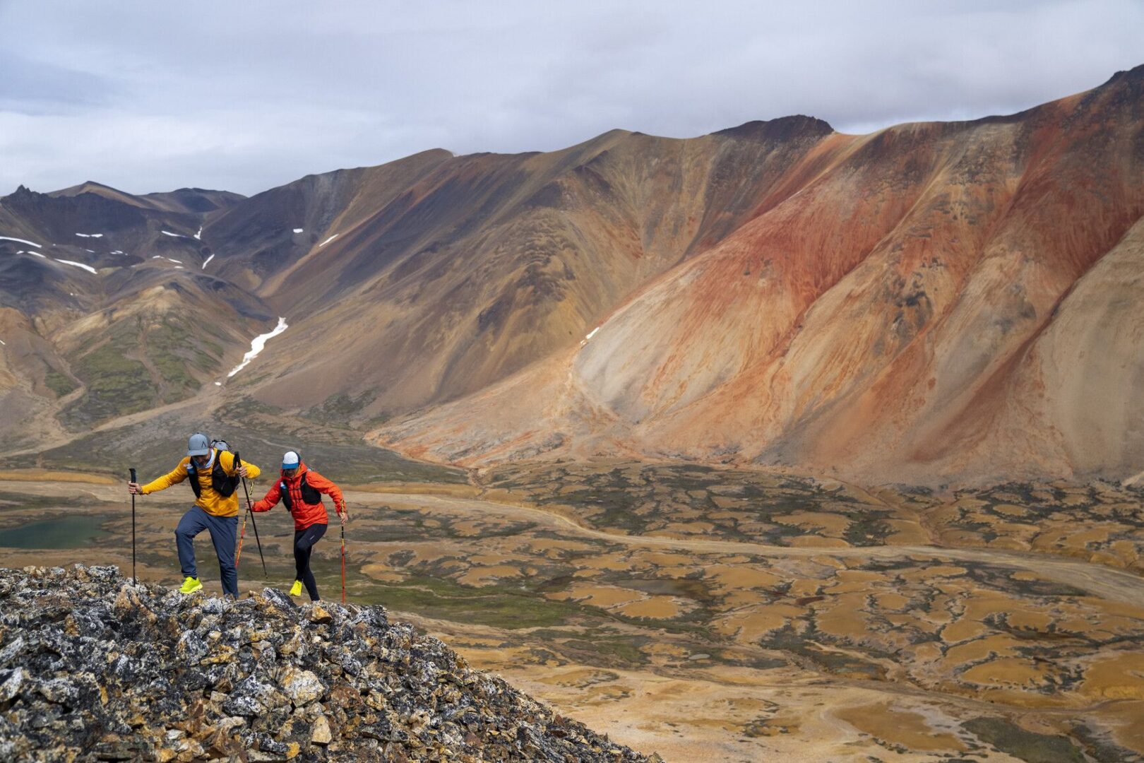Two hikers in bright jackets climb a rocky ridge with multicolored mountains behind them in a vast alpine landscape