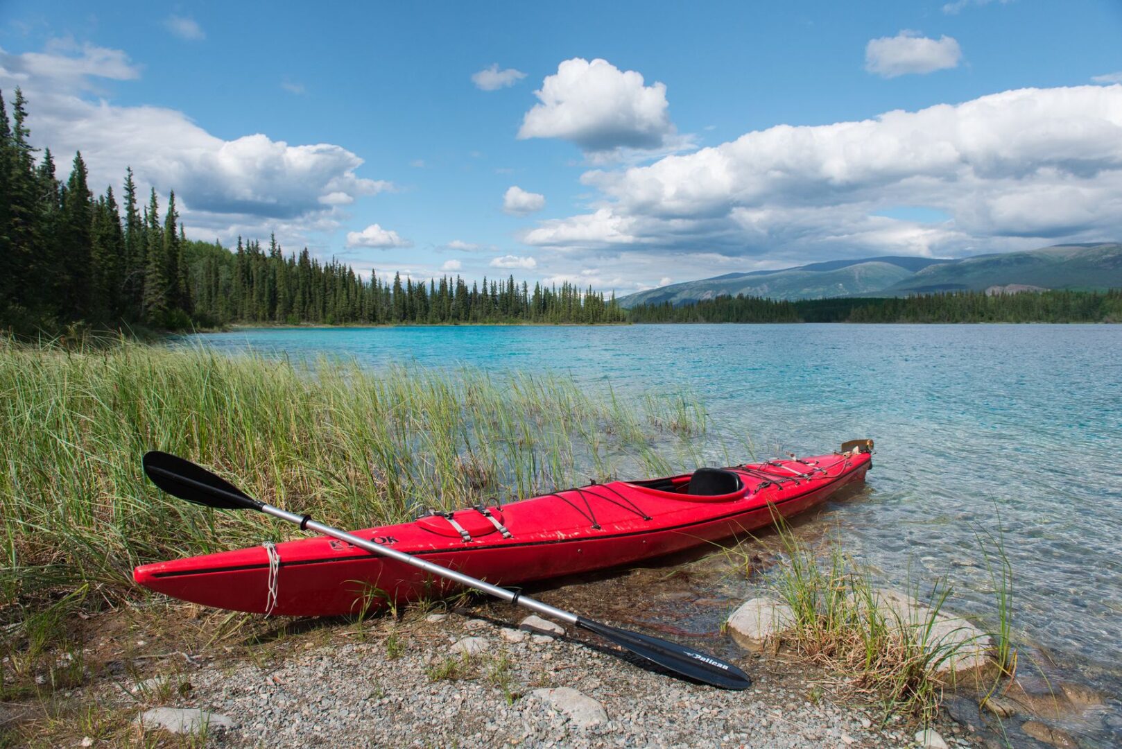 Red kayak resting on a rocky shore beside a clear turquoise lake with pine forest and distant mountains