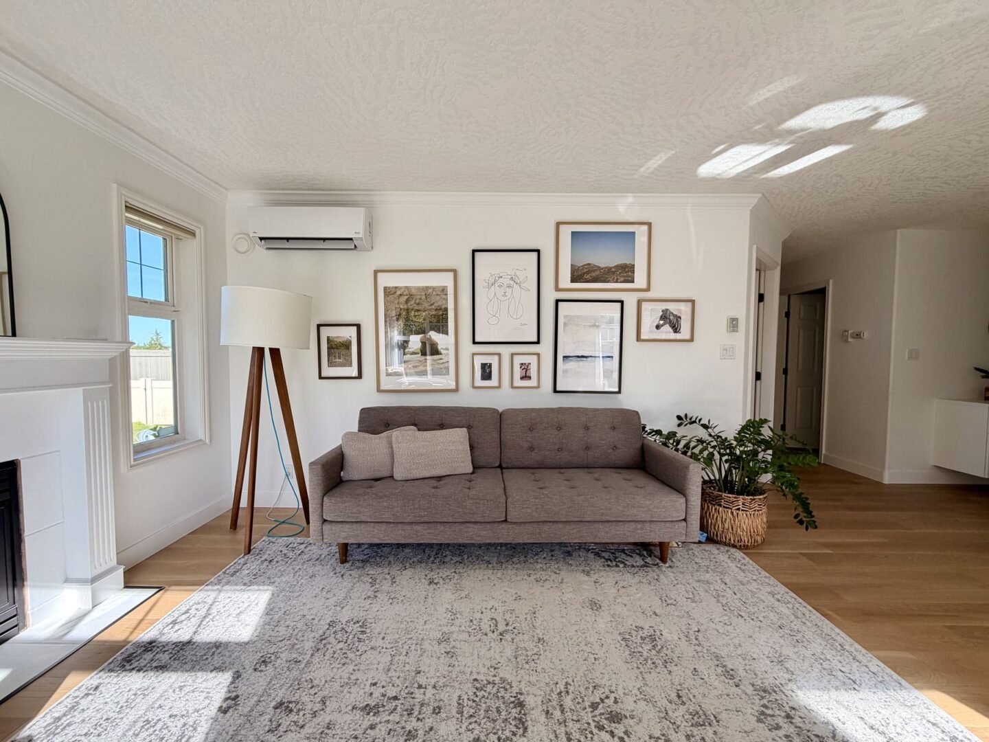 Bright living room with a gray tufted sofa gallery wall of framed art a wooden tripod floor lamp window on the left and a woven plant basket in the corner