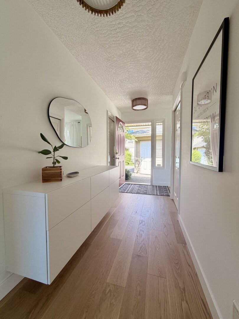 Bright modern hallway with white dresser round mirror and a small plant leading to an open front door showing a sunlit entrance