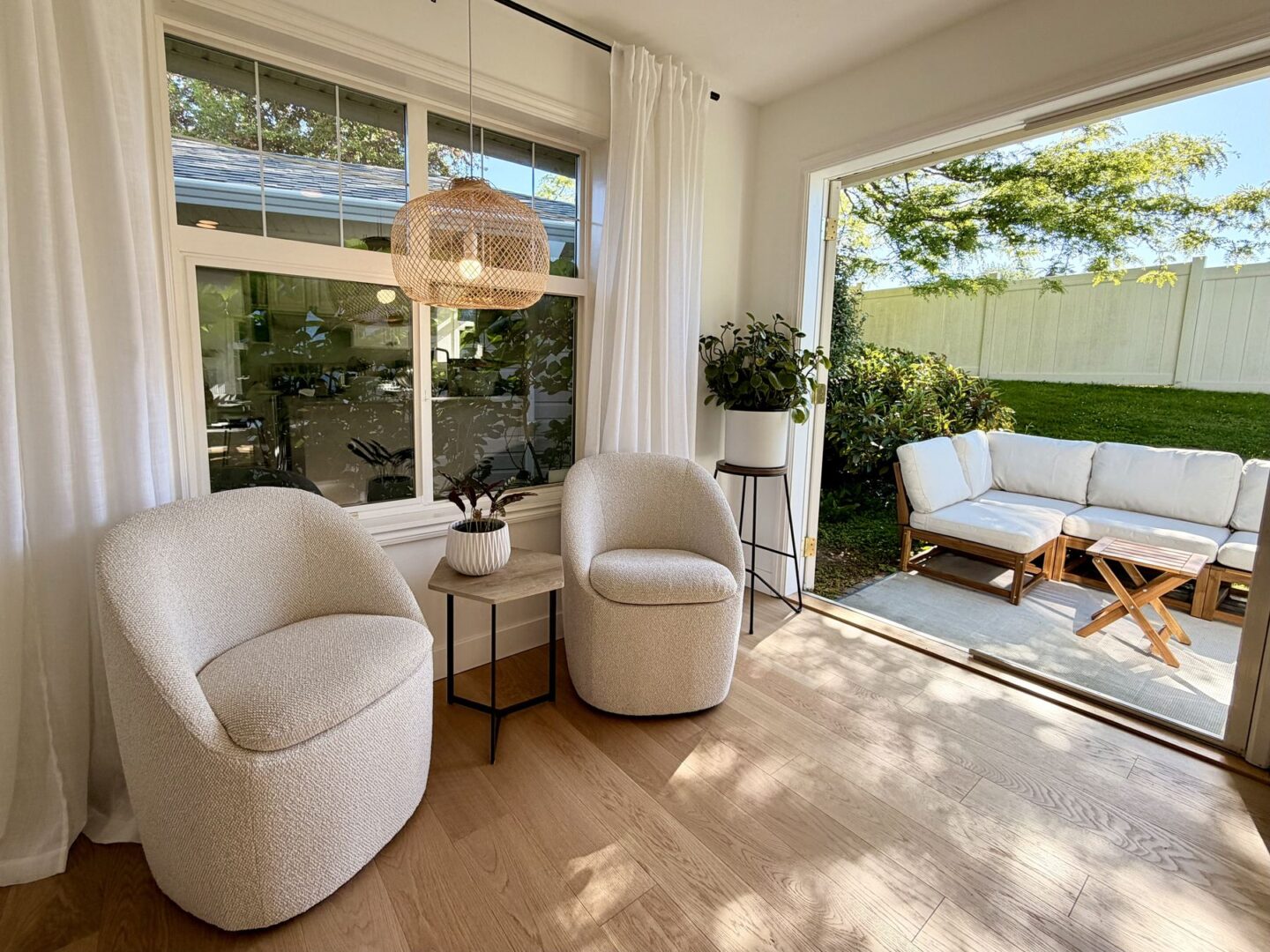 Cozy sunlit indoor seating area with two cream boucle chairs a small side table potted plant and a woven pendant light open doors lead to an outdoor white sofa