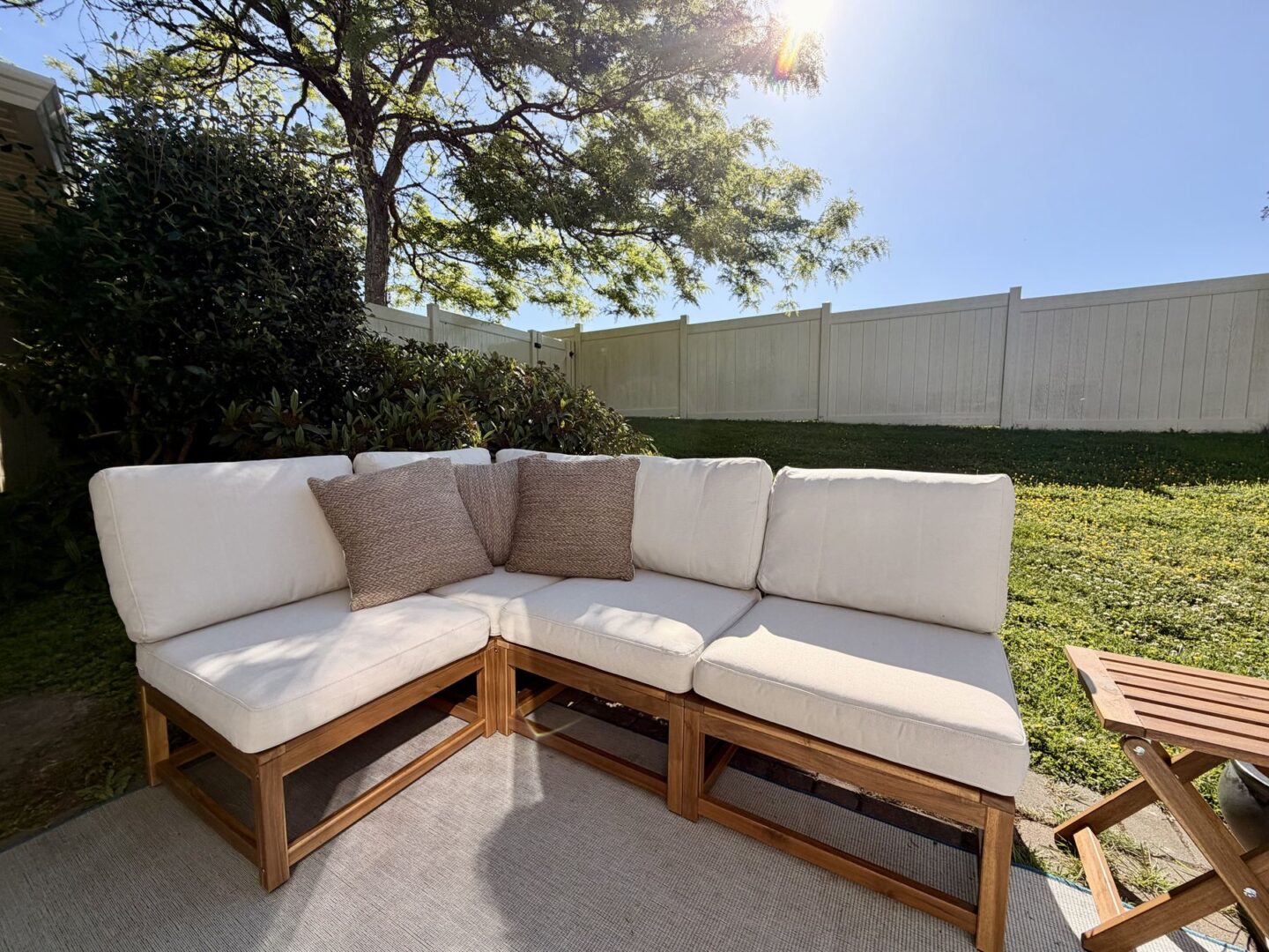 Sunlit backyard with a white cushioned outdoor sectional and beige throw pillows on a wooden frame facing a white fence and green lawn
