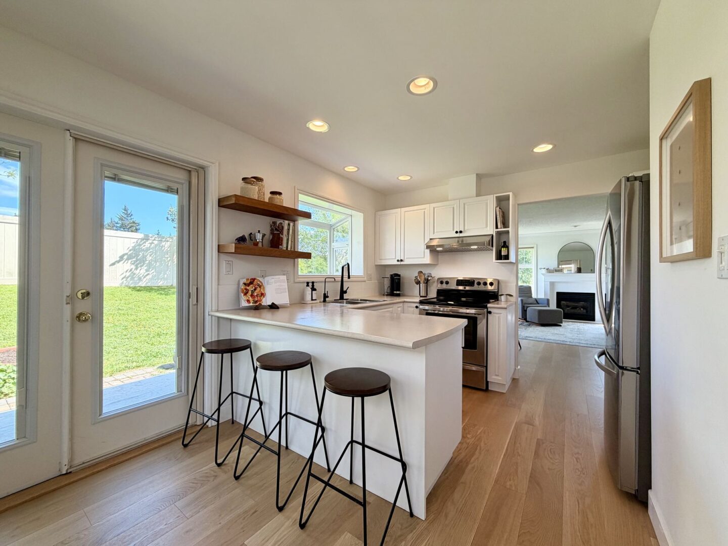 Bright white kitchen with an island and four black framed stools opening to a living room with a fireplace visible beyond the space