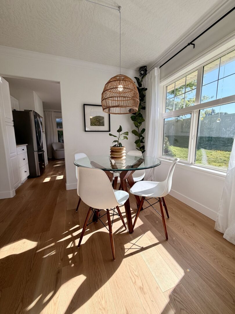 Sunlit dining nook with a glass round table white chairs a large wicker pendant light and tall green plants by a window
