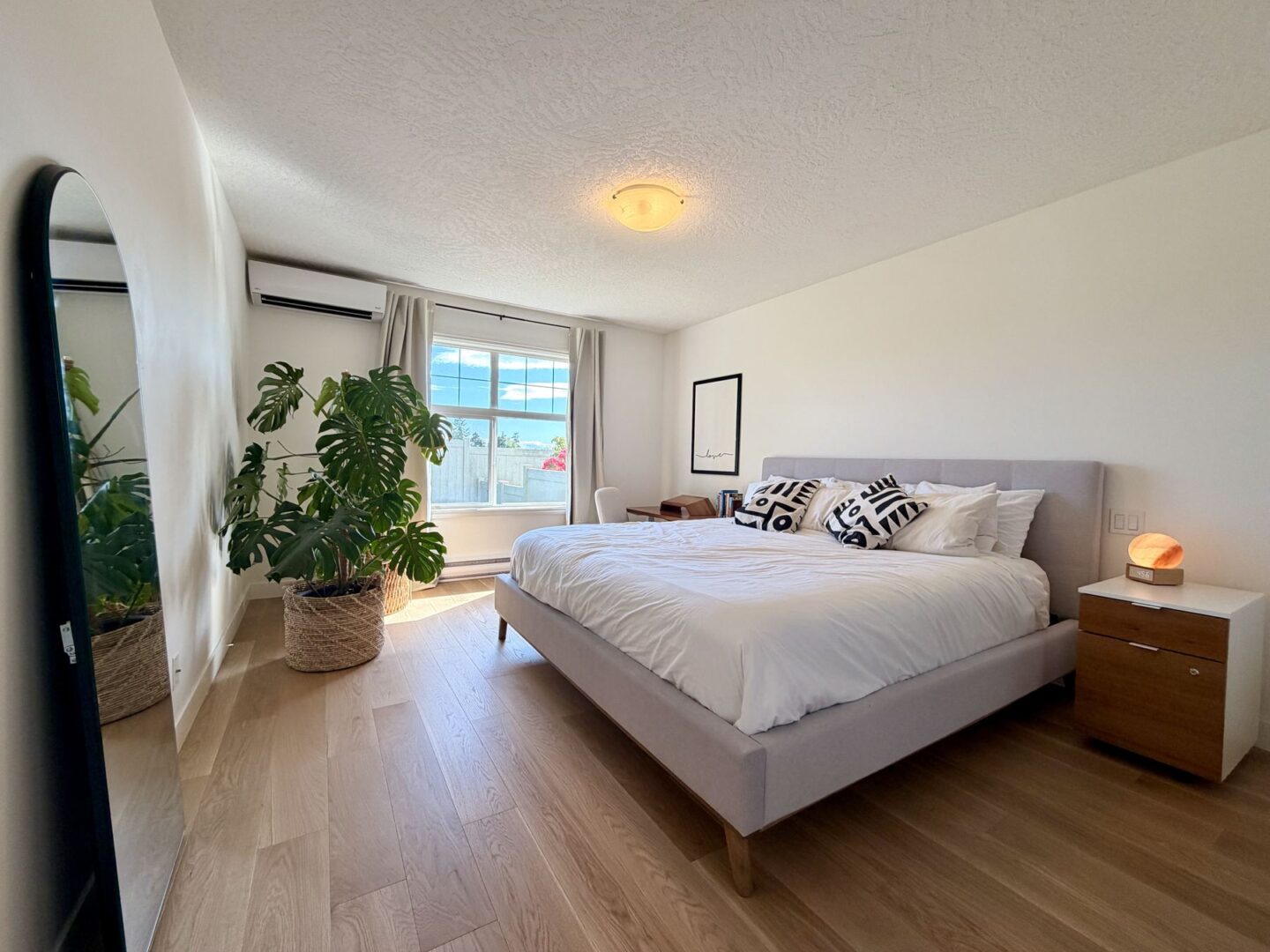 Bright modern bedroom with a gray upholstered bed white linens two wooden nightstands a framed print and a large monstera plant by a window