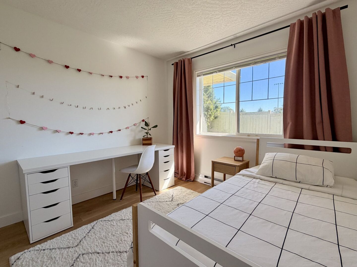 Bedroom with a white desk and drawers heart garland on the wall and a white bed by a window with pink curtains