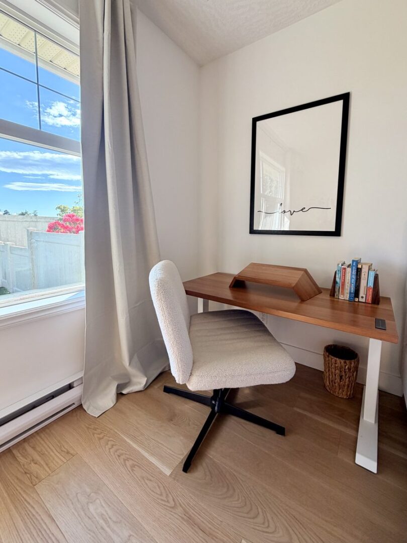 Cozy home office corner wooden desk with a curved bookrest cream swivel chair large window with gray curtains black framed art reading love books and a wicker wastebasket