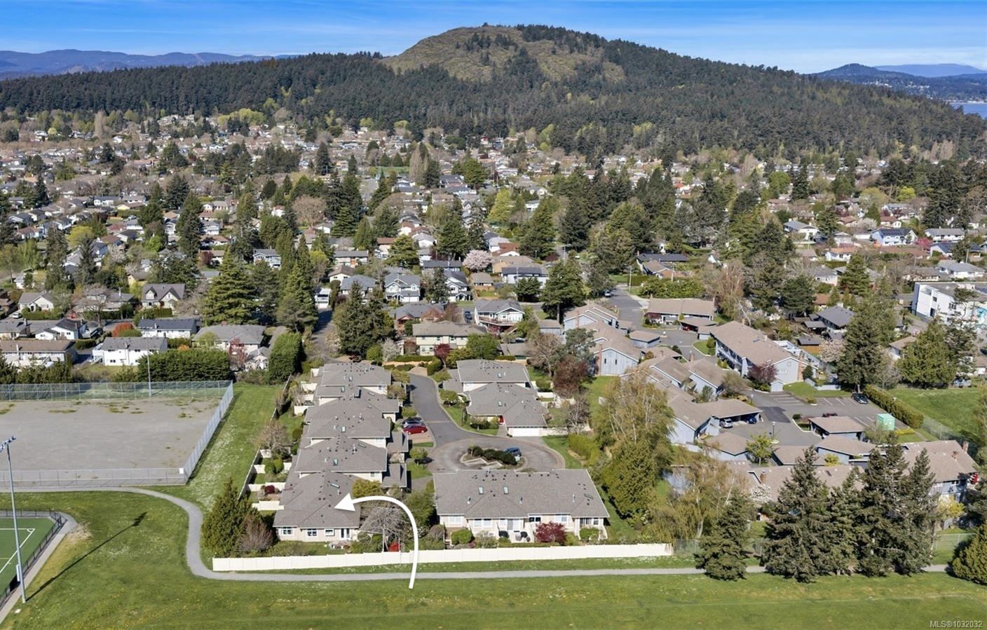 Aerial view of a suburban neighborhood with a white fenced house highlighted by a curved white arrow resting along a grassy field near a sports area and trees hills in the distance