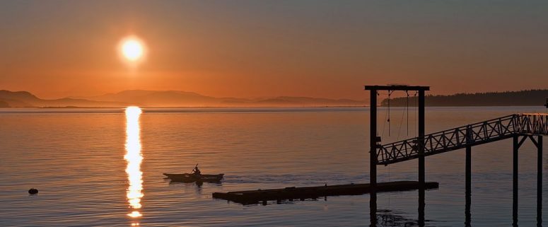 Sunset over calm water with a lone rowboat near a wooden dock and a bright sun reflecting on the surface