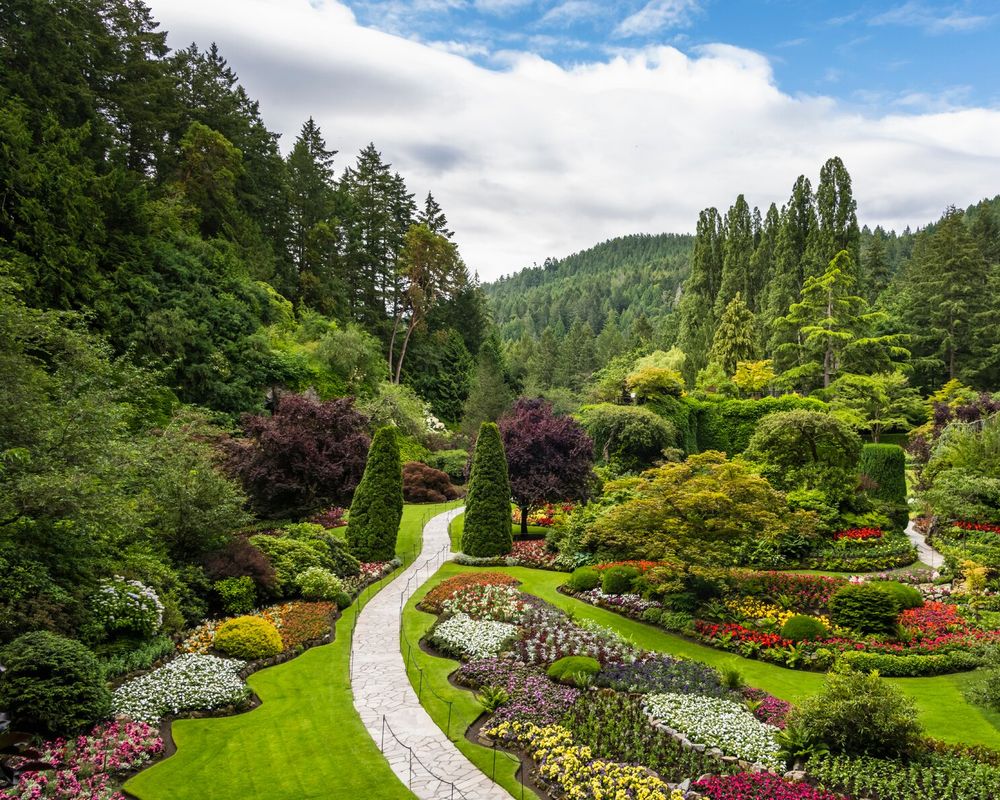 A winding stone path through a lush colorful formal garden bordered by vibrant flower beds and trimmed trees