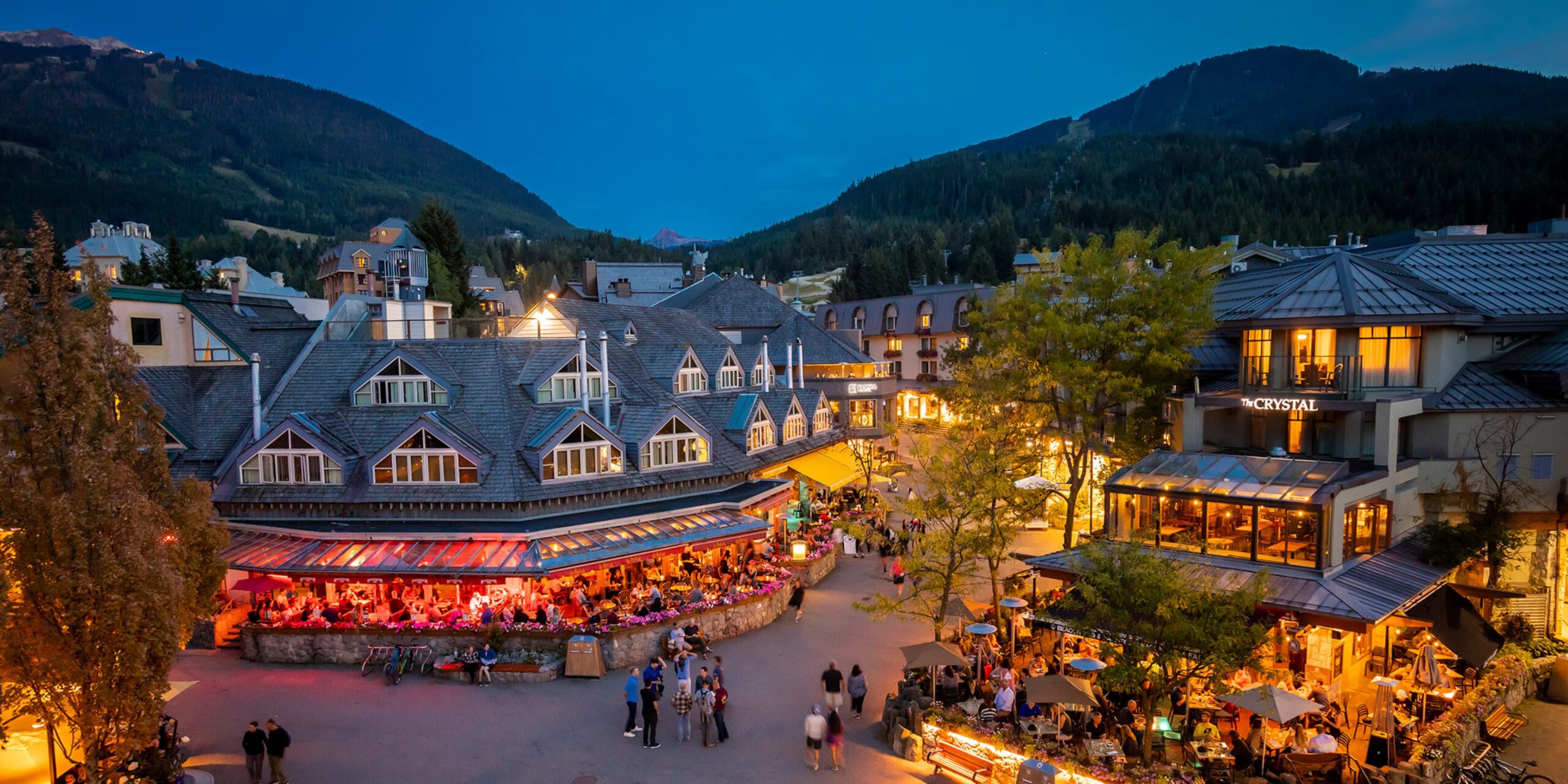 Alpine village square at dusk with outdoor restaurants lit by warm lights and people dining mountains in the background