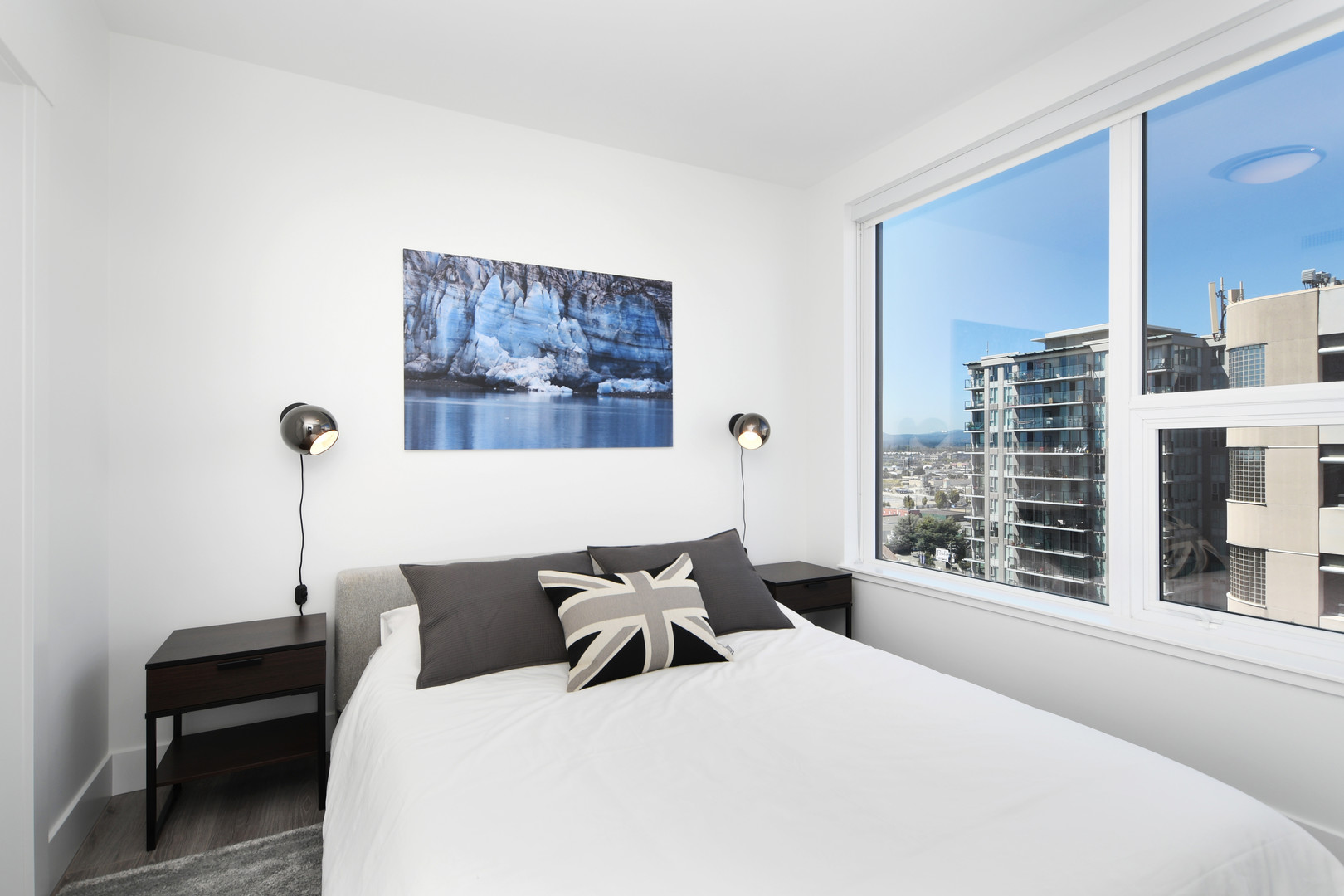 110644 | Apartments Homes Suites for Rent Victoria BC Modern bedroom with a white bed gray pillows Union Jack cushion two black nightstands and a large window showing high rise buildings outside