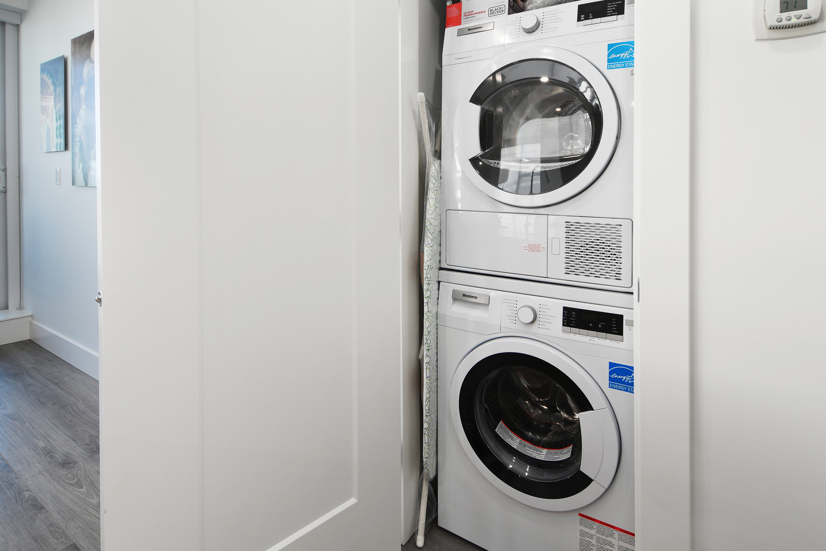 110648 | Apartments Homes Suites for Rent Victoria BC Stacked white front loading washer and dryer in a narrow laundry closet with the door open and a hallway nearby