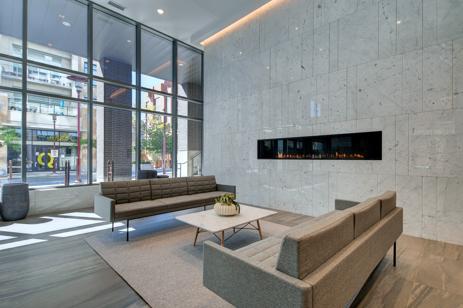 110652 | Apartments Homes Suites for Rent Victoria BC Modern lobby seating area with gray couches a white marble coffee table and a long linear fireplace set in a marble wall large windows show the city outside