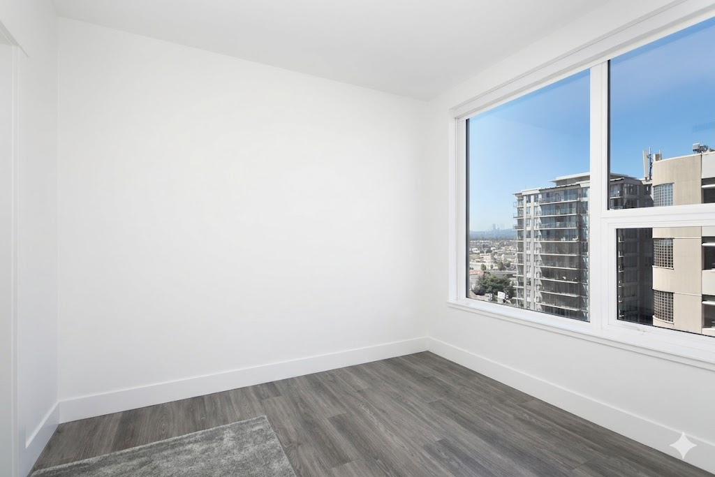 1106bedroom1 | Apartments Homes Suites for Rent Victoria BC Empty white room with a large window overlooking a cityscape gray wood like flooring and a small rug in the corner