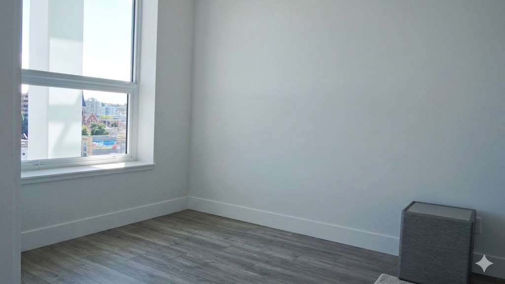 1106bedroom2 | Apartments Homes Suites for Rent Victoria BC Empty modern room with a large window showing city buildings light gray walls and gray wood laminate flooring a small gray storage box sits in the corner