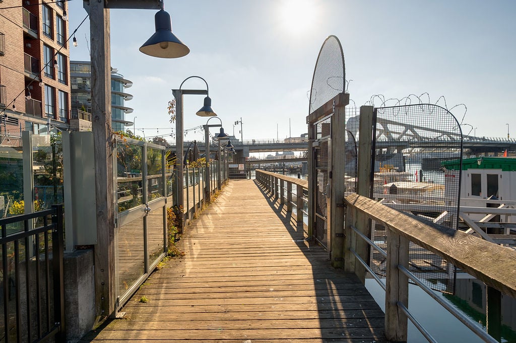 Sunlit wooden boardwalk along a harbor with glass railings on the left and a barbed wire fence on the right boats docked nearby