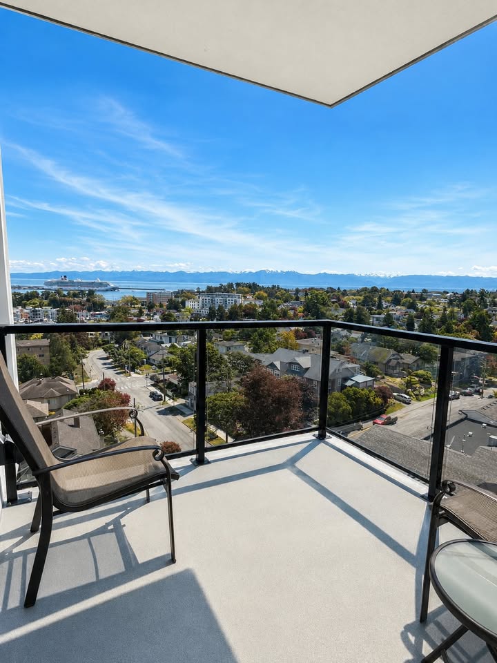 Balcony seating with two chairs and glass railing overlooking a suburban neighborhood a bay and distant mountains under a clear blue sky