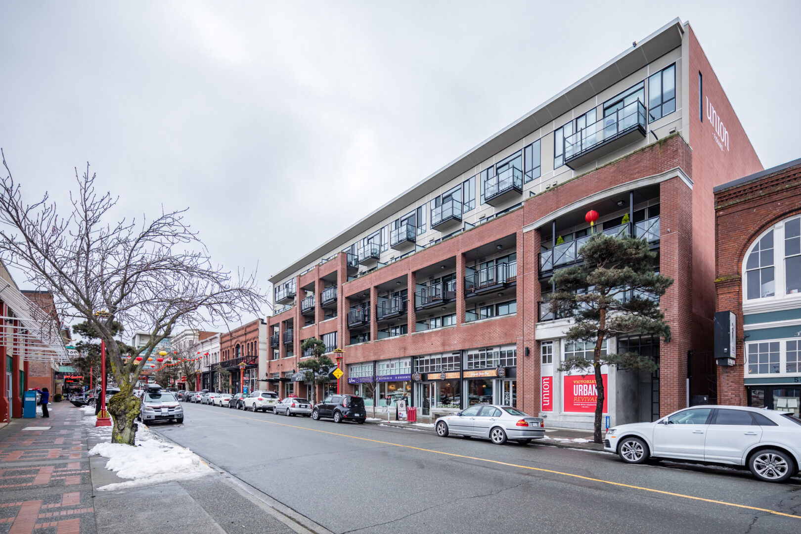 Urban street with a brick apartment building balconies storefronts parked cars and bare trees in winter