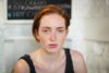 Portrait of a young woman with short red hair and freckled skin looking at the camera in a light colored room with a chalkboard in the background wearing a black sleeveless top